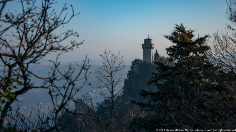Distant crenellated tower on a wooded rocky ridge, framed by bare branches against a hazy horizon.