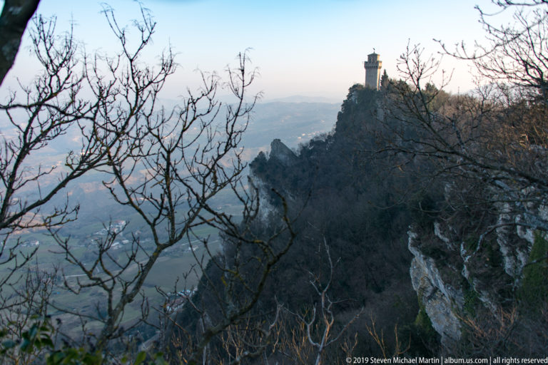 Distant fortress tower on a steep wooded ridge above pale cliffs, framed by bare branches and hazy landscape.