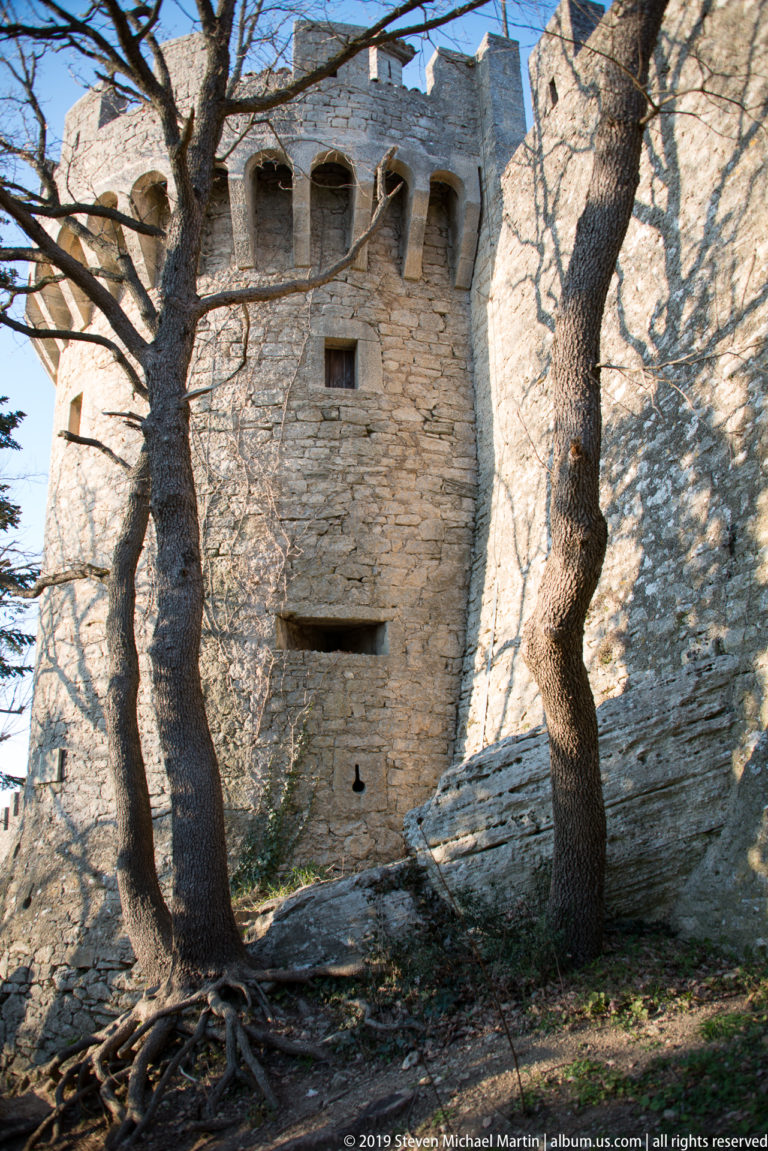 Square stone defensive tower with projecting upper supports, narrow openings, and trees growing close to its walls.