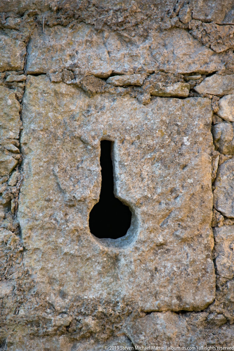 Small keyhole-shaped opening cut through a large rectangular stone block in rough fortress masonry.