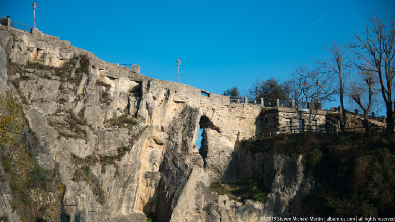 Low stone fortification crossing a rough rock face, with a narrow opening, parapet, bare trees, and blue sky.