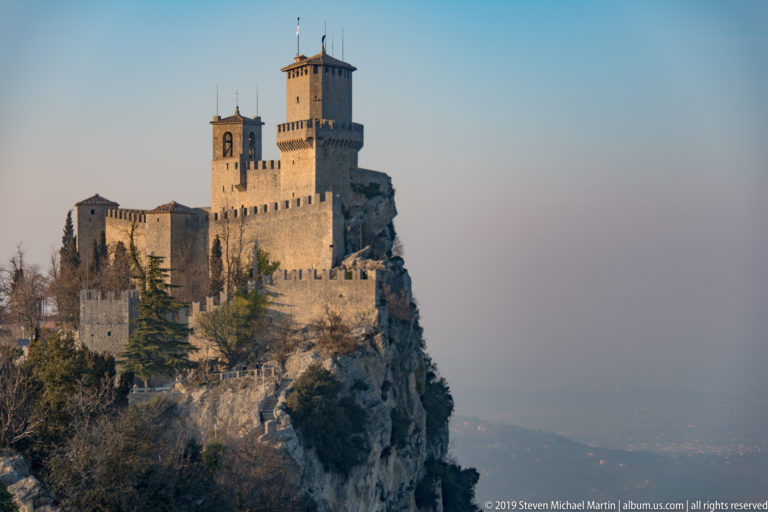 Guaita fortress filling a rocky promontory with tall keep, bell tower, crenellated walls, trees, and hazy landscape beyond.