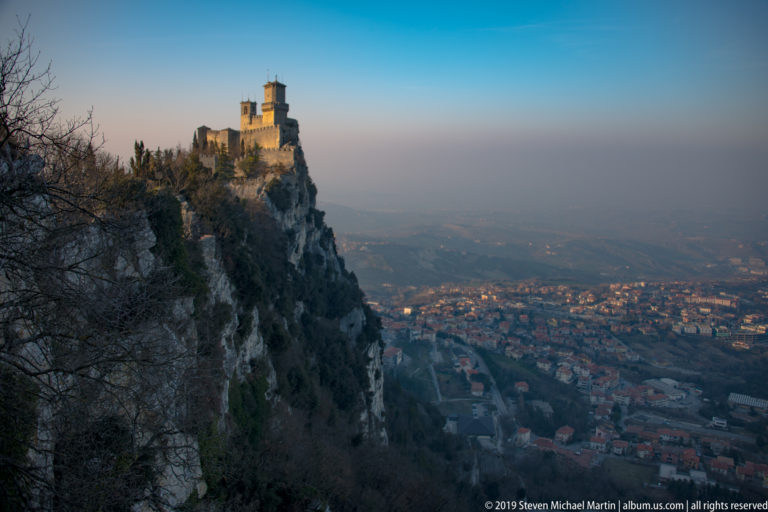 Distant Guaita fortress on a high cliff above a dense town and hazy valley, framed by bare branches.