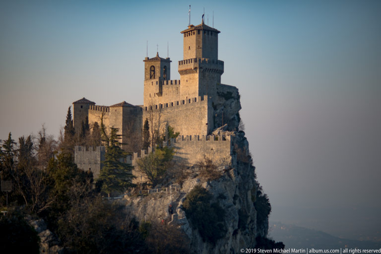 Guaita fortress with tall keep, bell tower, and crenellated walls perched on the sheer rocky edge of Monte Titano.