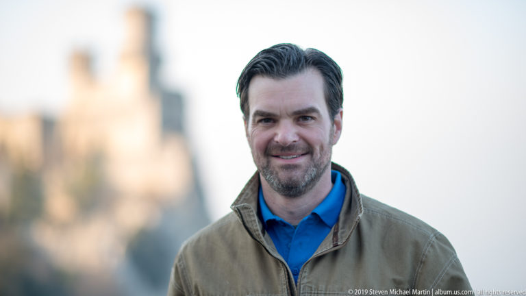 Bearded man in tan jacket and blue shirt posed before a strongly blurred hilltop fortress.