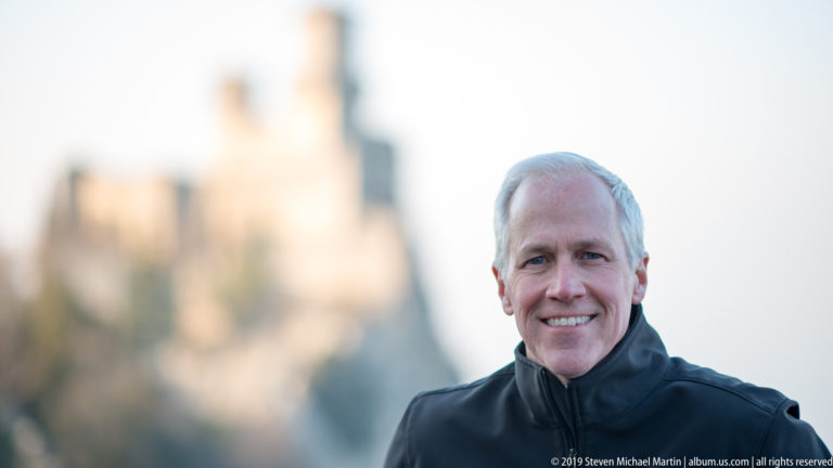 Smiling gray-haired man in a dark jacket sharply focused before a heavily blurred hilltop fortress.