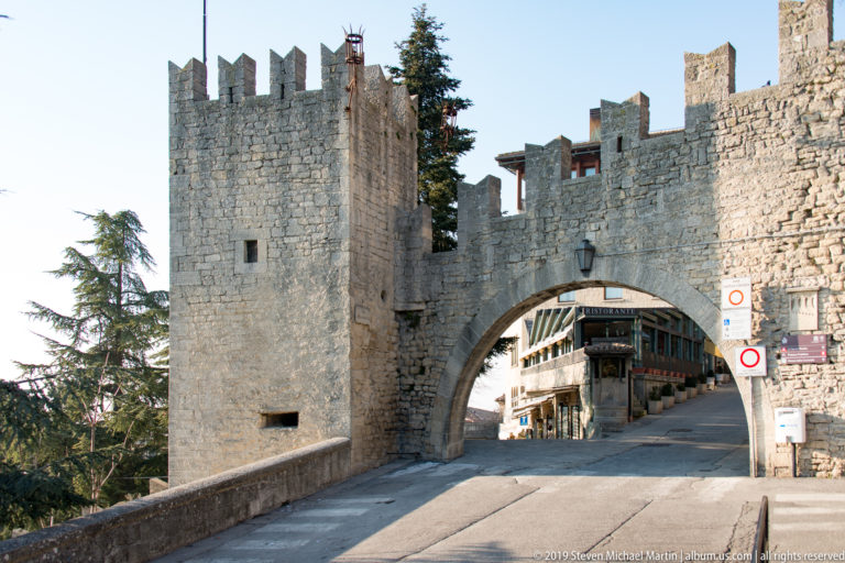 Square stone tower and crenellated wall flank a broad arched gateway leading to an uphill street in San Marino.