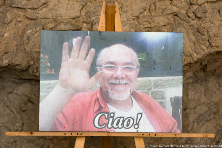 Printed portrait of a smiling man waving on a wooden easel against stone, with the word Ciao across the bottom.