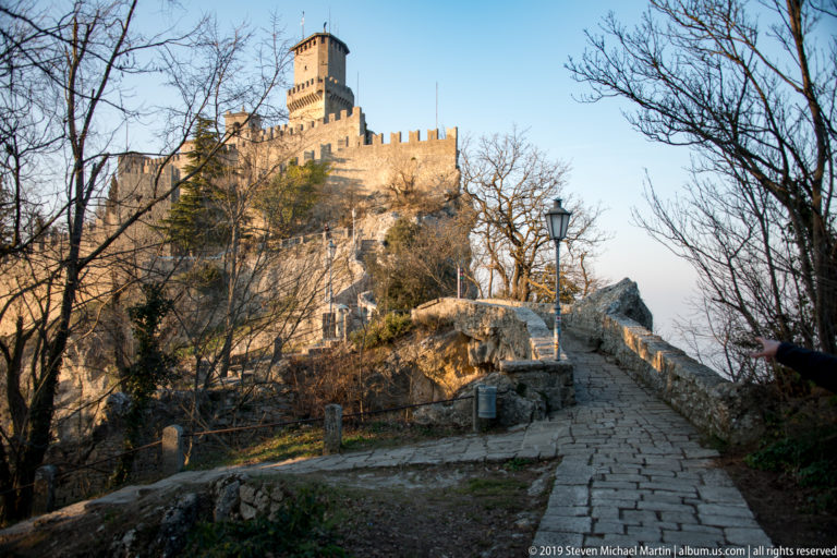 Stone walkway with low parapets and lamp ascending through leafless trees toward Guaita’s tall keep and crenellated walls.