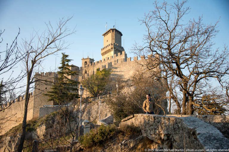 Guaita’s tall keep and crenellated walls above a rocky terrace, leafless trees, and a visitor standing on a low wall.