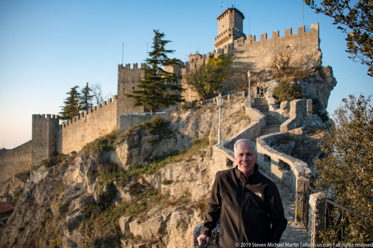 Visitor standing on a rocky path below the crenellated walls and tall keep of the First Tower, Guaita.