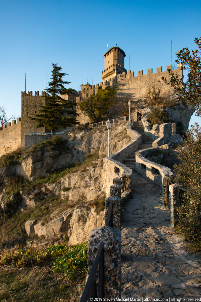 Stone path zigzagging over exposed rock toward the crenellated walls and tall keep of the First Tower, Guaita.