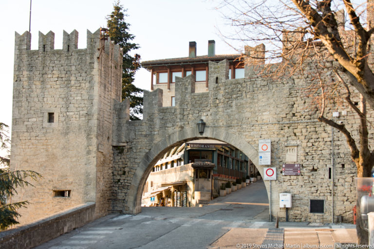 Large arched gateway through thick stone fortifications, with towers, traffic signs, and a street of shops visible beyond.