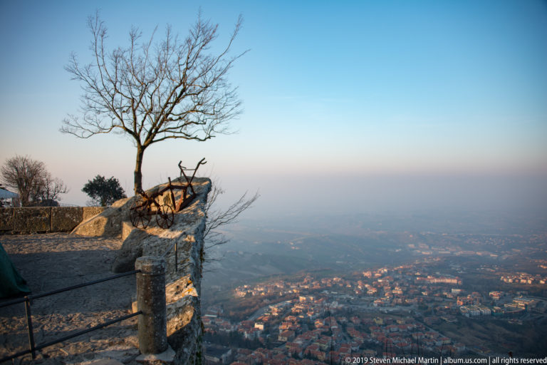 Leafless tree and exposed roots on a rocky overlook above dense neighborhoods and hazy countryside below Monte Titano.