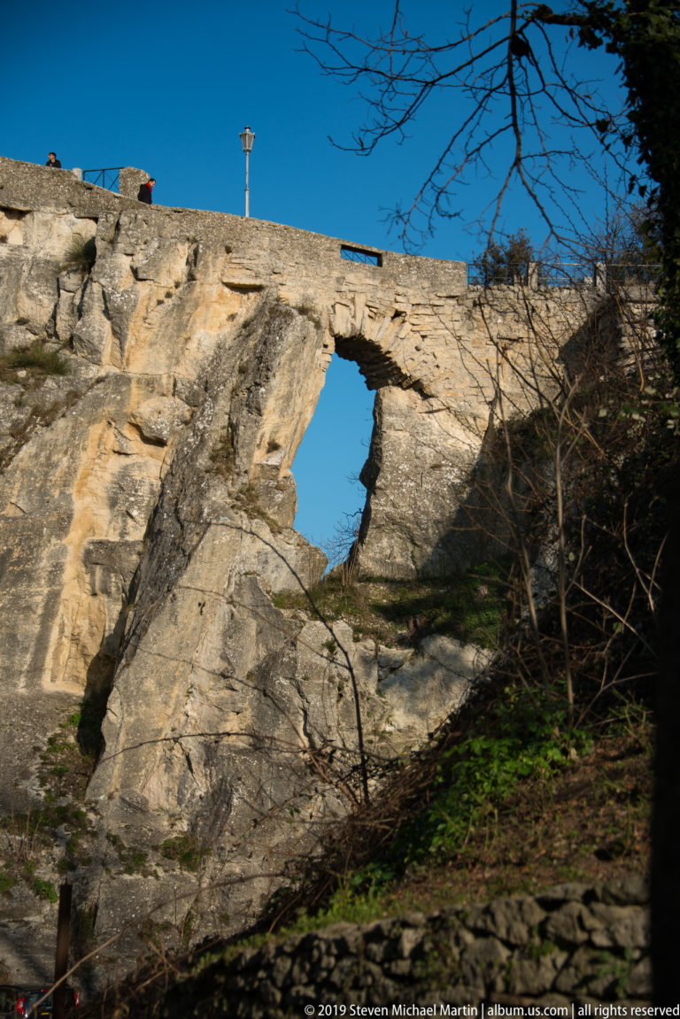 Irregular hole through pale weathered rock below a stone parapet, with bare branches and low vegetation around the slope.