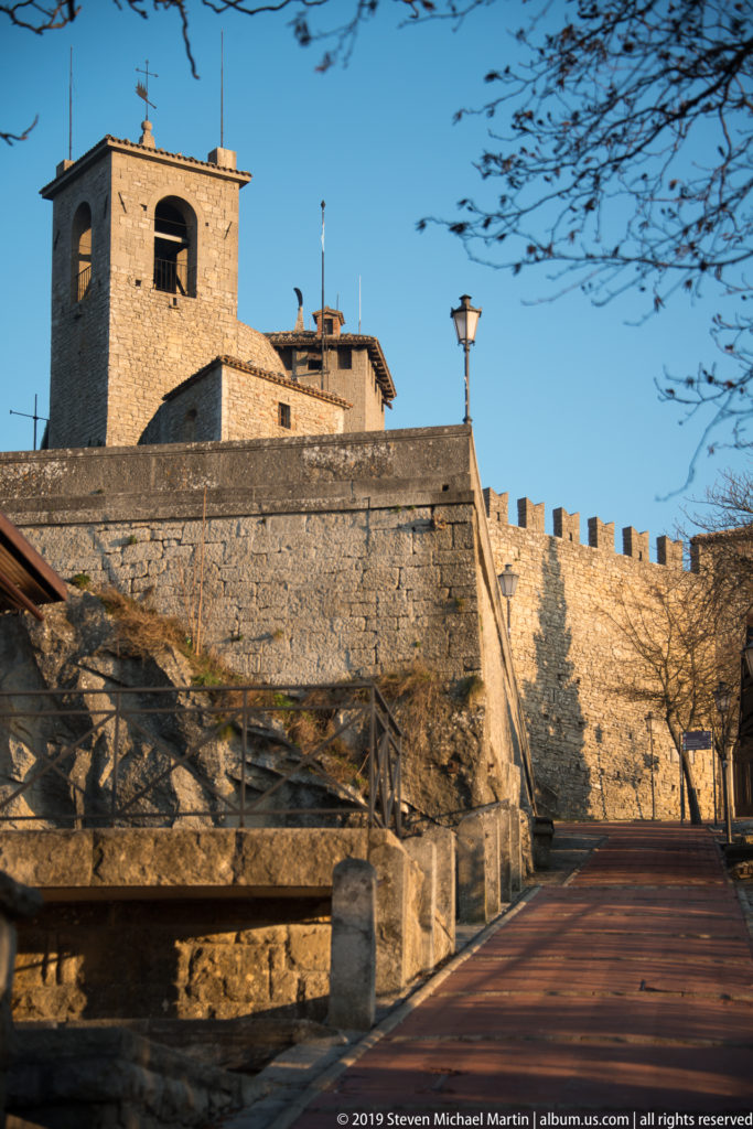 Ascending paved walkway below a crenellated stone wall and square bell tower, with rock terraces and bare branches.