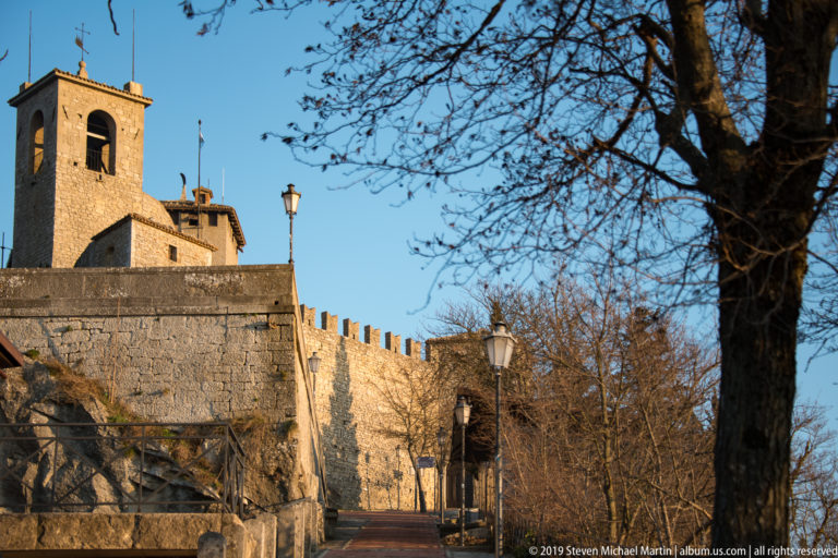 Crenellated stone wall and square bell tower beside a paved path lined with leafless trees and streetlamps.