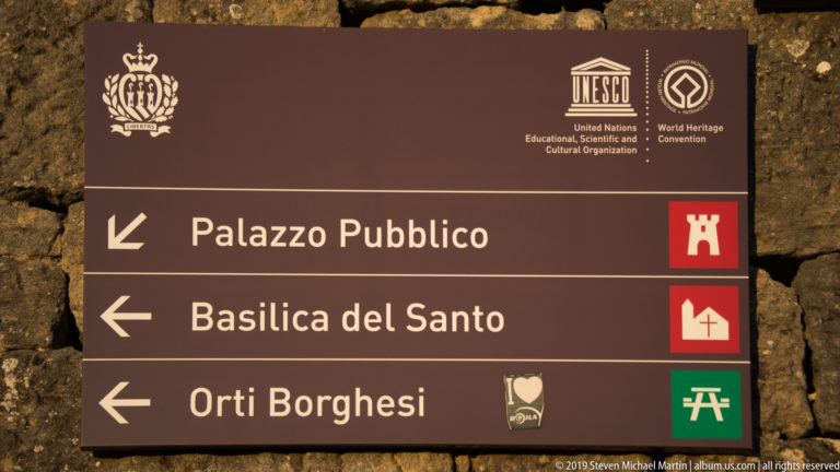 Brown UNESCO wayfinding sign pointing left to Palazzo Pubblico, Basilica del Santo, and Orti Borghesi.
