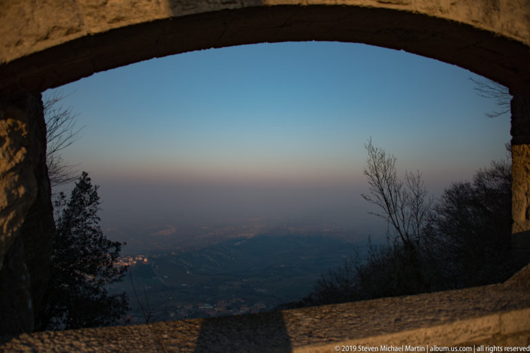 Large stone arch framing a hazy valley view with bare trees, low parapet, and distant settlements below Monte Titano.