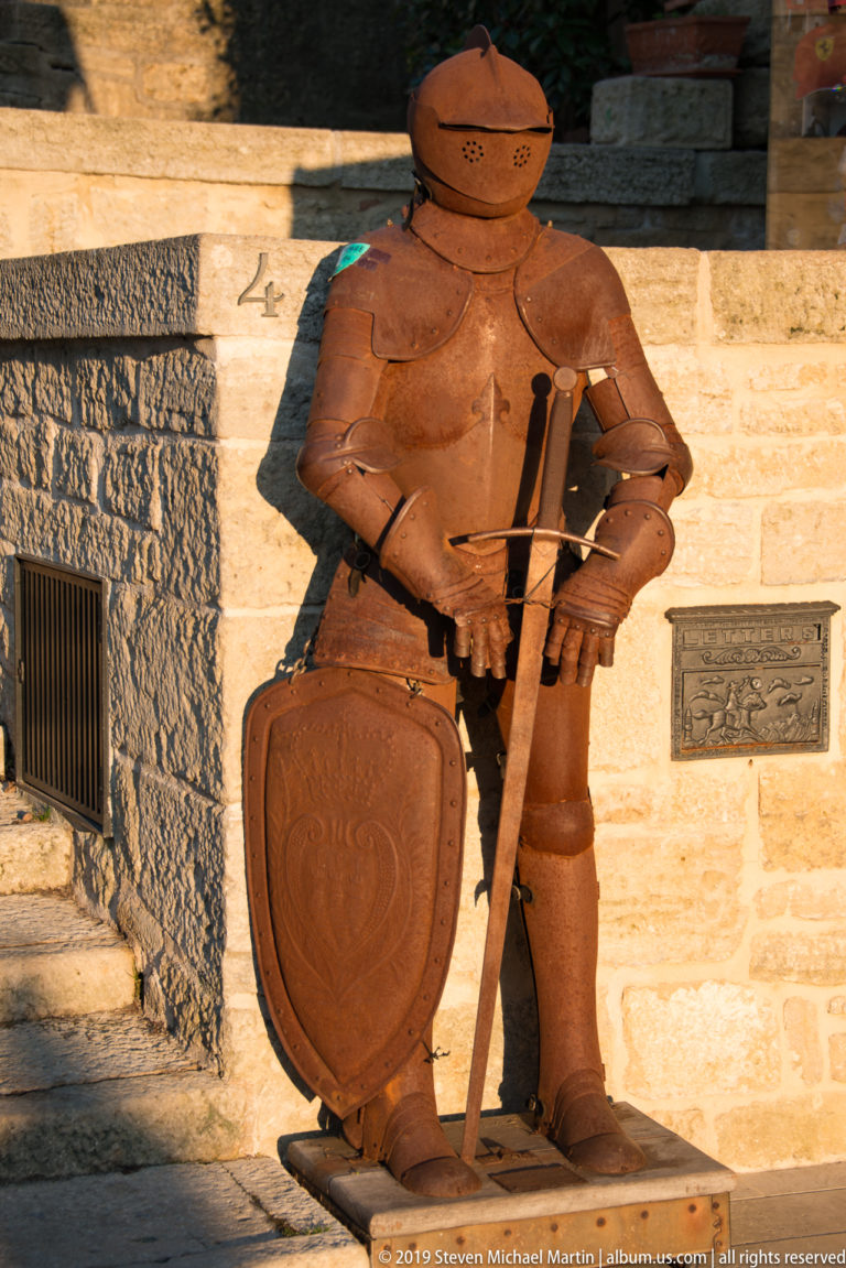 Rust-colored armored knight display holding a pole weapon and shield embossed with a crowned three-tower emblem.
