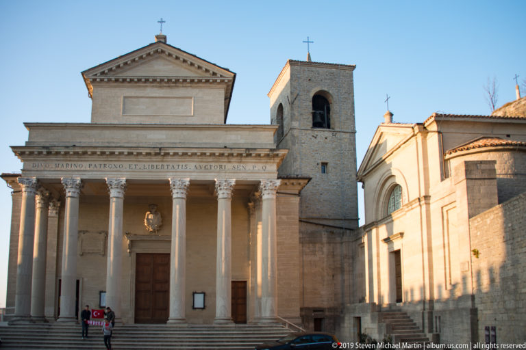 Basilica di San Marino with Corinthian portico and steps, square bell tower, and smaller adjoining church under a clear sky.