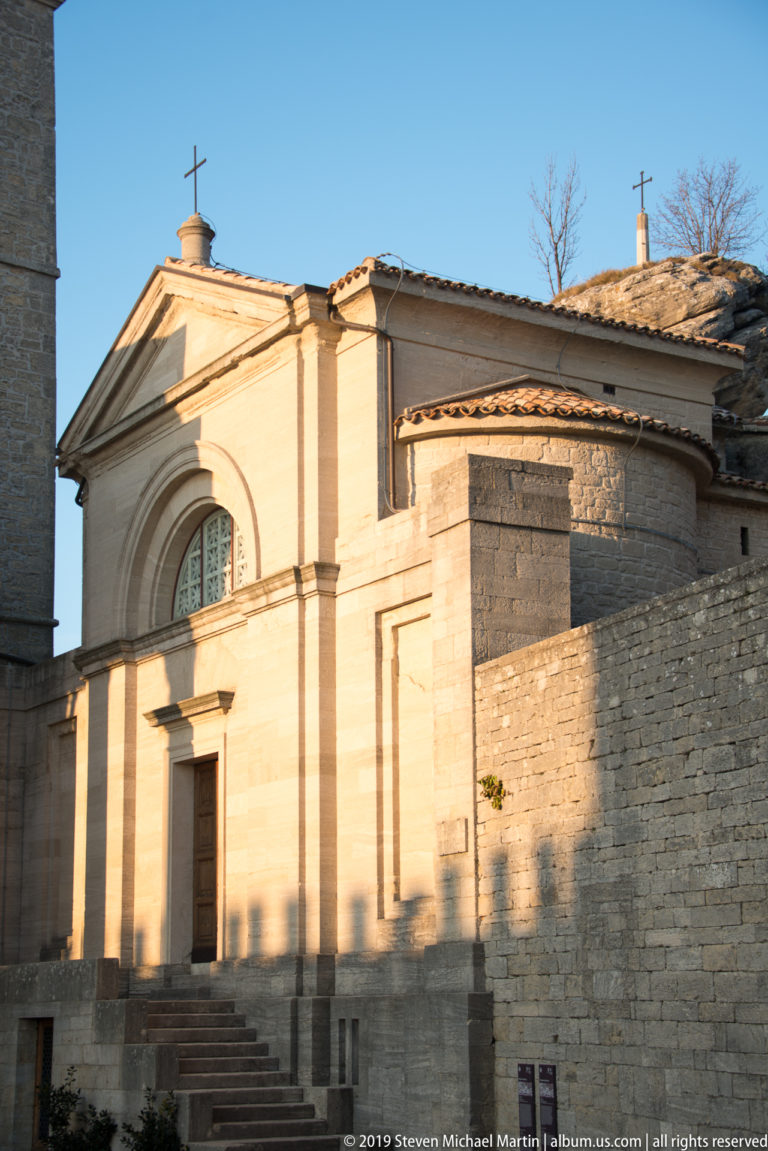 Oblique view of the Small Church of Saint Peter with pedimented façade, arched window, tiled roofs, and adjoining stone structures.