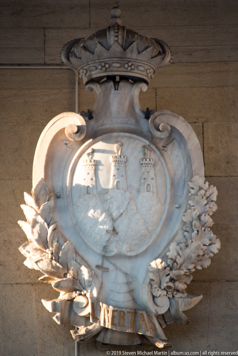 Carved stone San Marino coat of arms with three towers, crown, leafy branches, and LIBERTAS motto.
