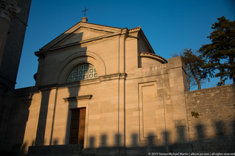Small Church of Saint Peter beside the Basilica of Saint Marinus, with pediment, arched window, central doorway, and roof cross.