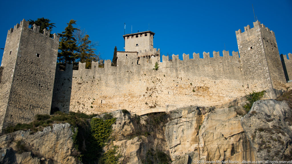 Large crenellated stone fortress with multiple square towers rising above a steep rocky slope under blue sky.