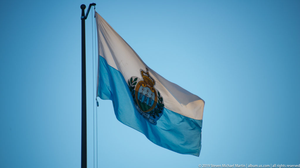 White and light-blue San Marino flag flying from a pole with the crowned national coat of arms at center.