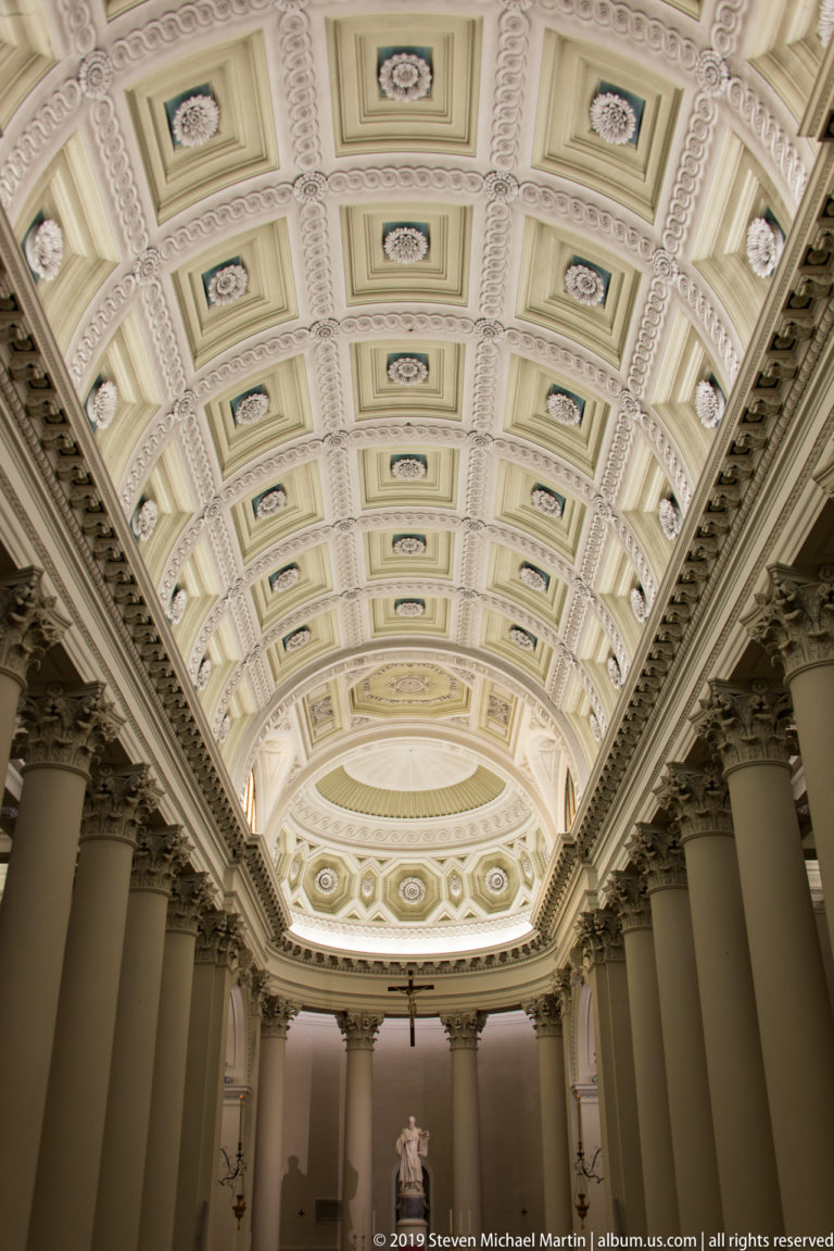 Ornate coffered basilica ceiling with rosettes and pale green accents extending between column rows toward the sanctuary dome.