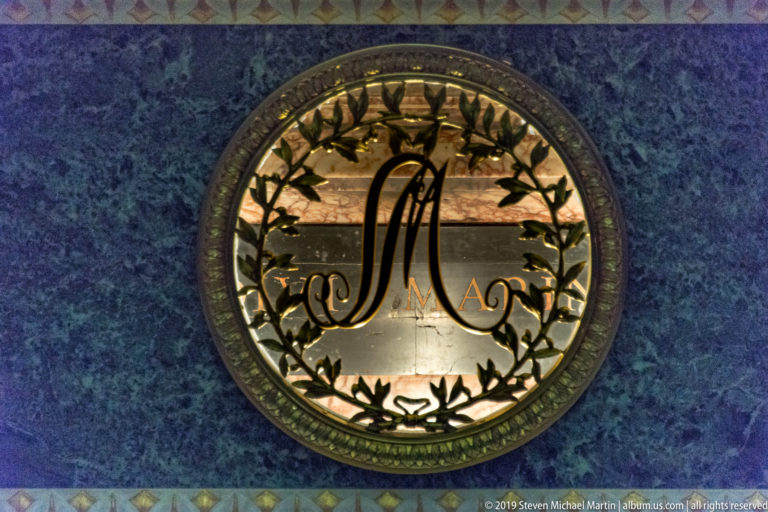 Circular metalwork medallion with a large scripted M surrounded by gilded leaves on a blue-green marble background.