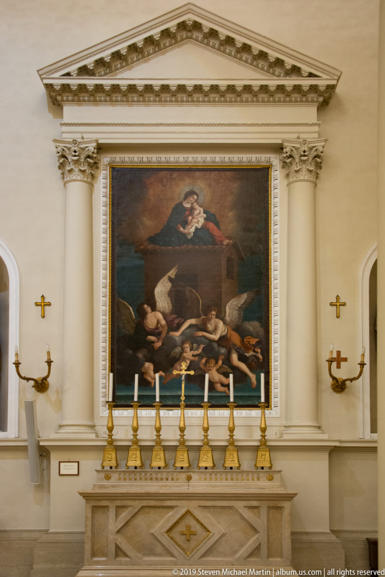 Classical side altar with a large religious painting, paired columns, triangular pediment, and a row of gold candlesticks.