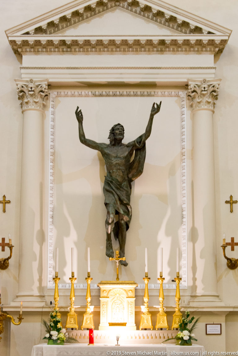 Large bronze-toned male saint with raised arms above an altar lined with gold candlesticks, flowers, and devotional lights.