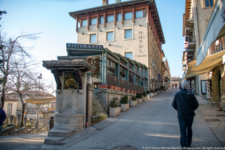 Uphill stone-lined street with restaurant frontage, Hotel Bellavista signage, a small roofed monument, and one pedestrian.
