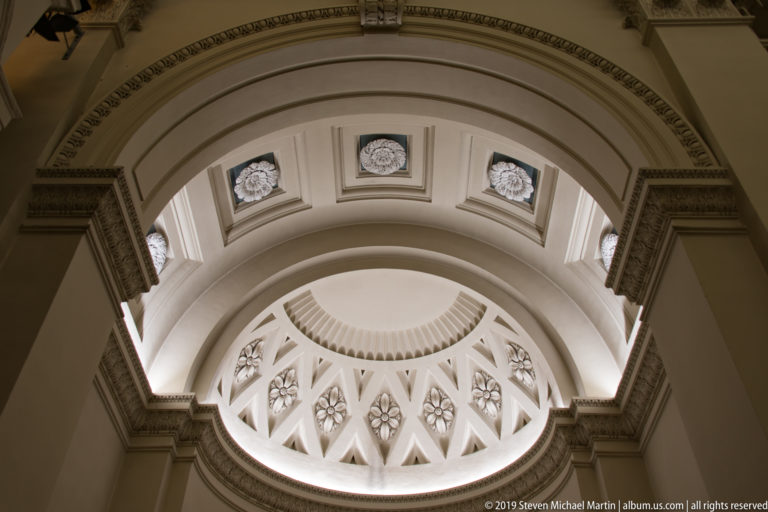 Upward view of layered cream arches, recessed panels, rosette windows, and geometric tracery beneath a semicircular basilica vault.