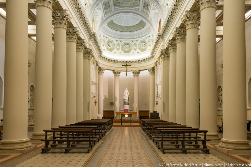 Centered view down Basilica di San Marino nave with tall columns, wooden benches, patterned aisle, ornate ceiling, and sanctuary.