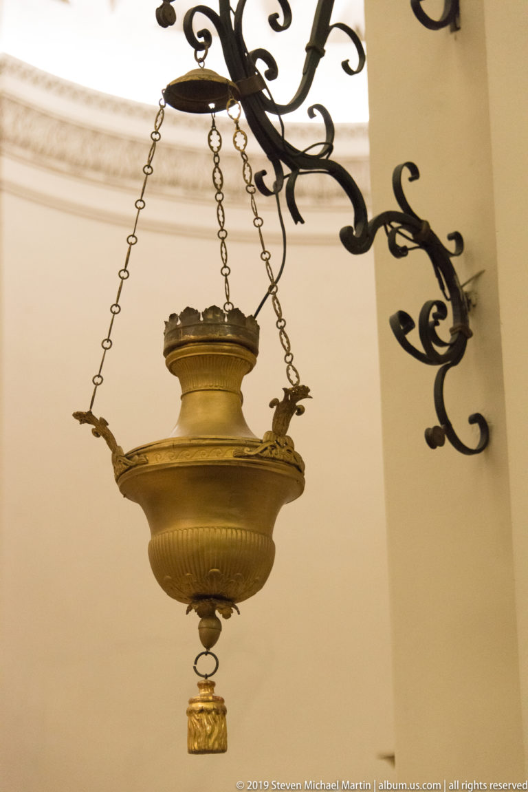 Large brass hanging sanctuary lamp suspended by chains from an ornate black wrought-iron wall bracket.