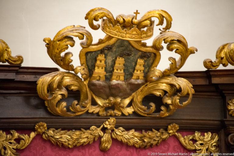 Gilded carved cartouche with San Marino’s three-tower shield, crown, cross, scrolling foliage, and cherubic face.