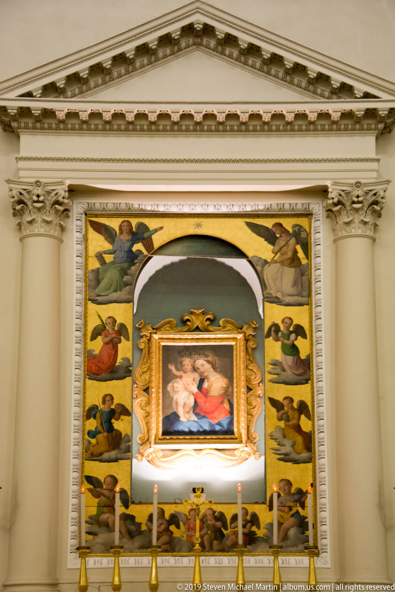 Side altar with a small framed Virgin and Child painting surrounded by painted angels, classical columns, pediment, and candles.