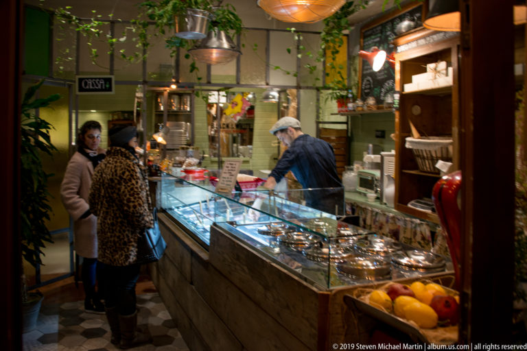 Two customers and a shop worker at a glass delicatessen counter filled with prepared foods.