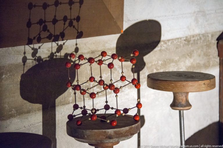 Red-and-dark molecular model on a round pedestal casting a large shadow beside a narrow table.