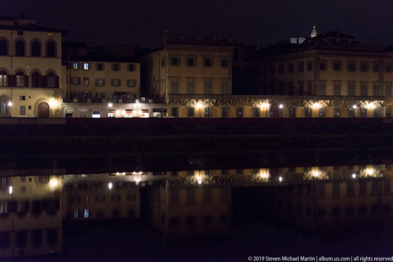 Illuminated historic buildings along the Arno reflected in dark, calm water at night.