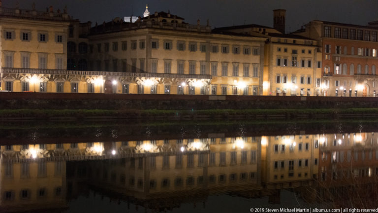 Long row of illuminated Florentine palazzi and embankment reflected in the Arno at night.