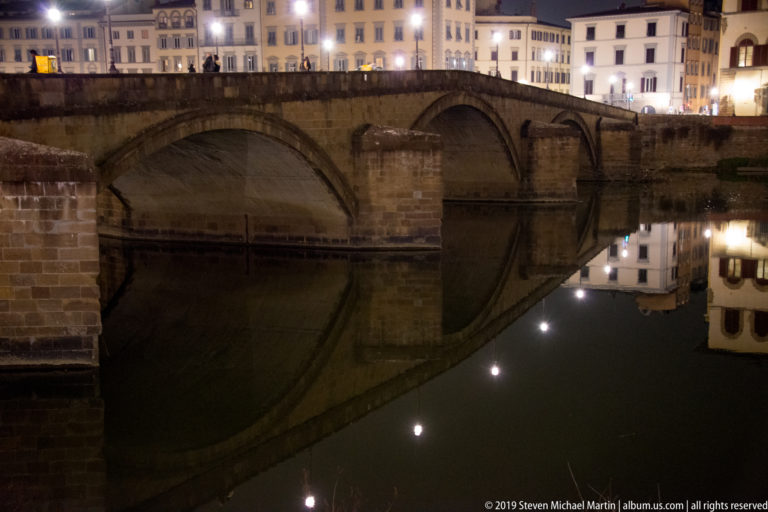 Three-arched Ponte Santa Trinita crossing the Arno at night, with bridge and illuminated buildings reflected in still water.
