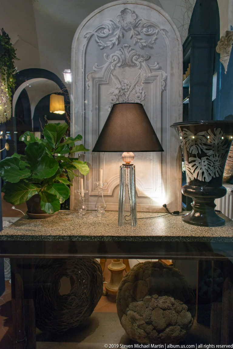 Black-shaded table lamp, leafy plant, carved pale relief panel, and decorative vessels arranged in a shop interior.