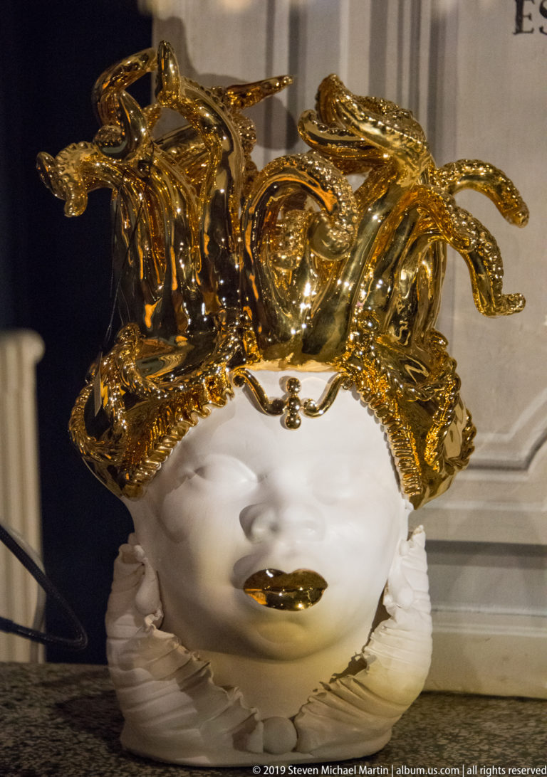 White decorative face mask with gold lips and an elaborate crown of curling gilded ornament displayed indoors.