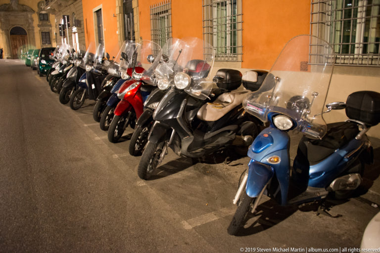 Long row of parked motor scooters beside an orange building wall, with a blue scooter closest to the camera.