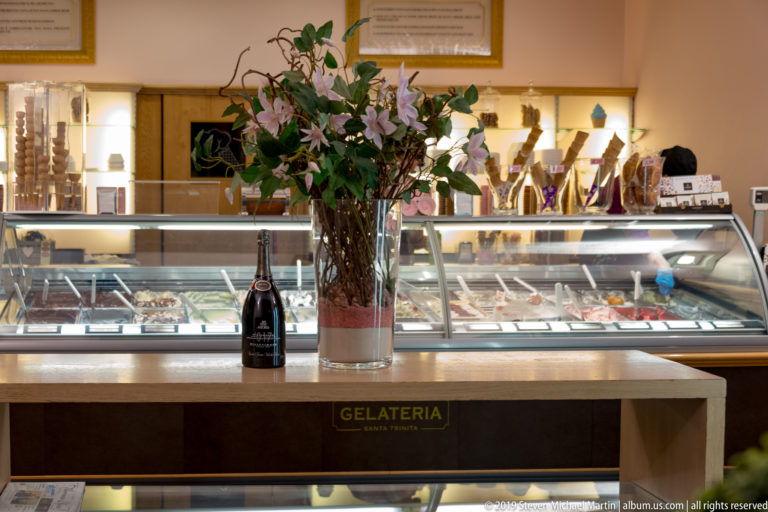 Gelateria counter with glass dessert case, vase of lilies and greenery, and a bottle under warm interior lighting.