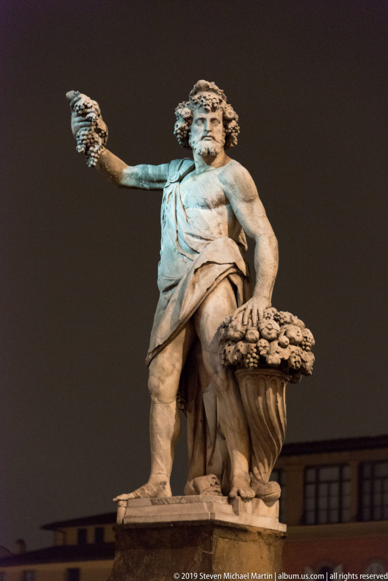 Illuminated stone male figure with drapery holding a small vessel above an ornate fountain basin at night.